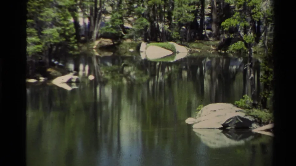 A handheld 8mm view of Lake Tahoe shows calm water, reflected pine trees, and rounded granite rocks along the wooded shore.