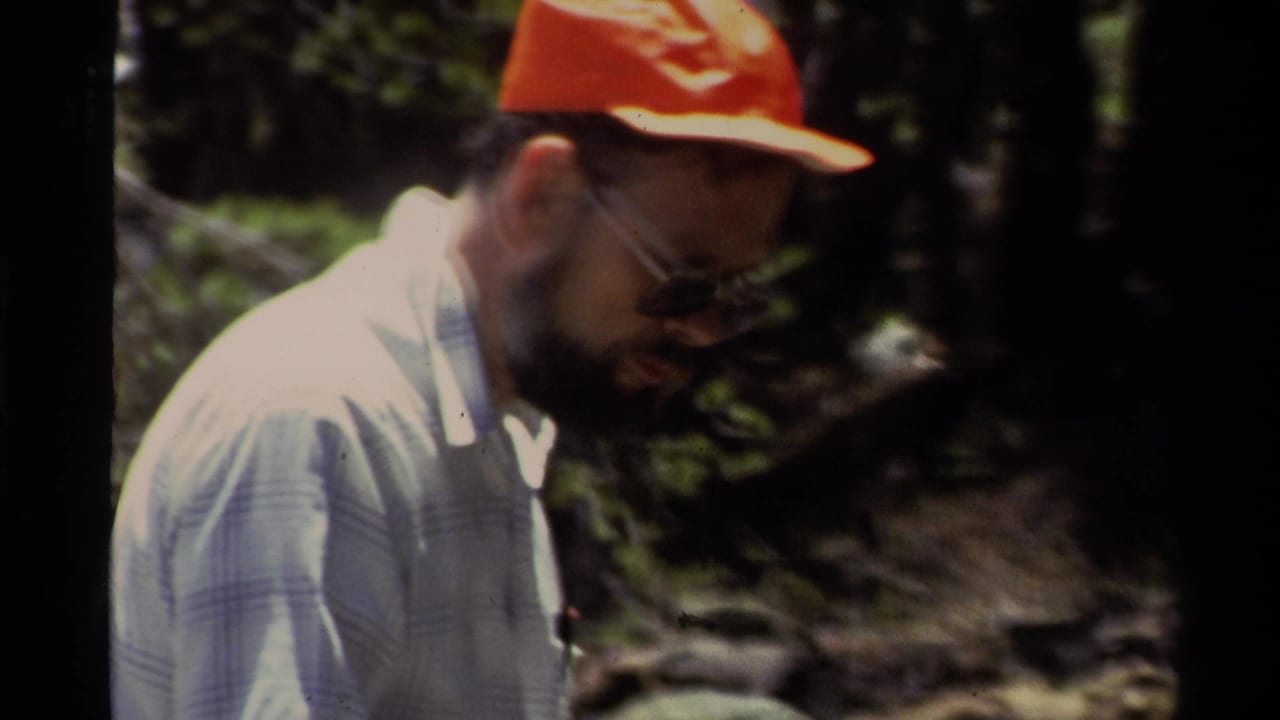 In Lake Tahoe home-movie footage from 1978, a man in a red cap and glasses looks down near trees and rocky ground.