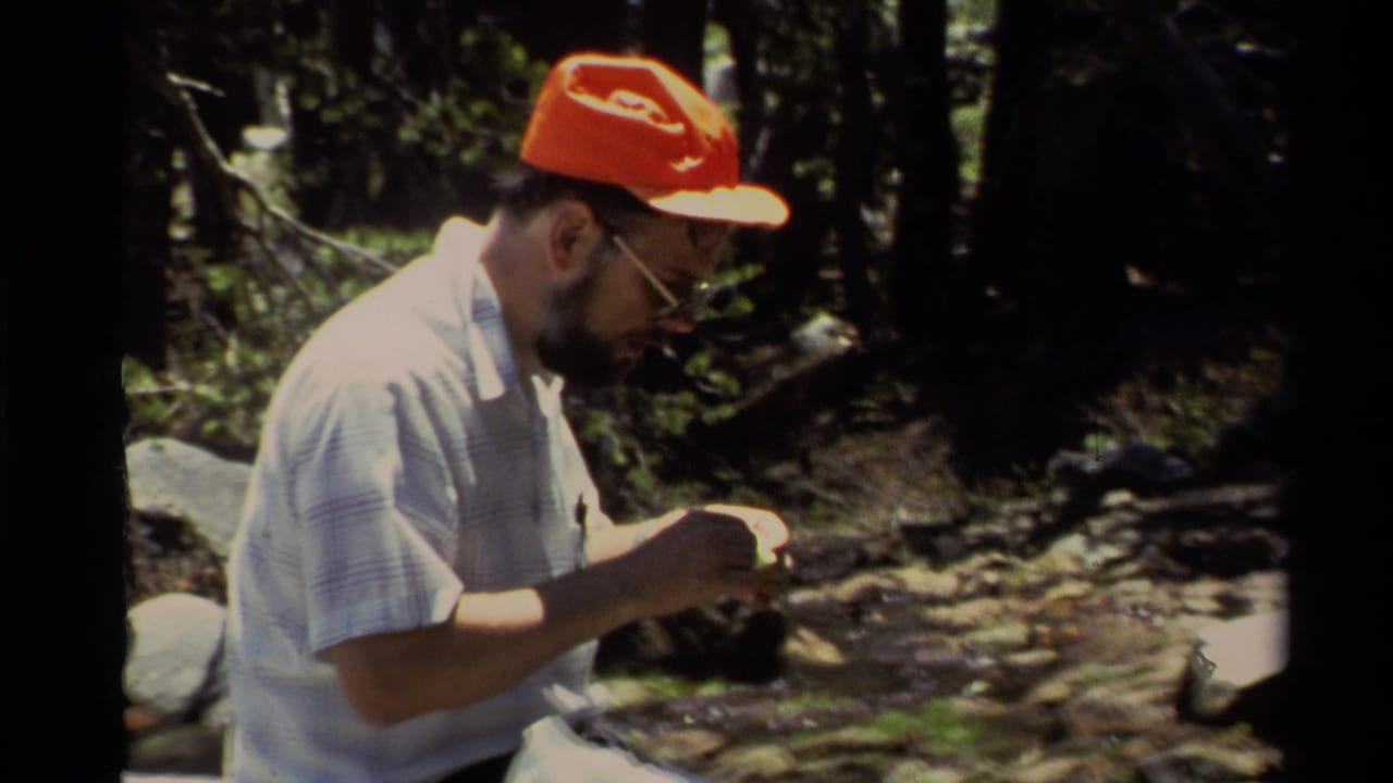 The man sits by a rocky creek or shoreline in 1978 Lake Tahoe footage, focused on fishing line or tackle in his hands.
