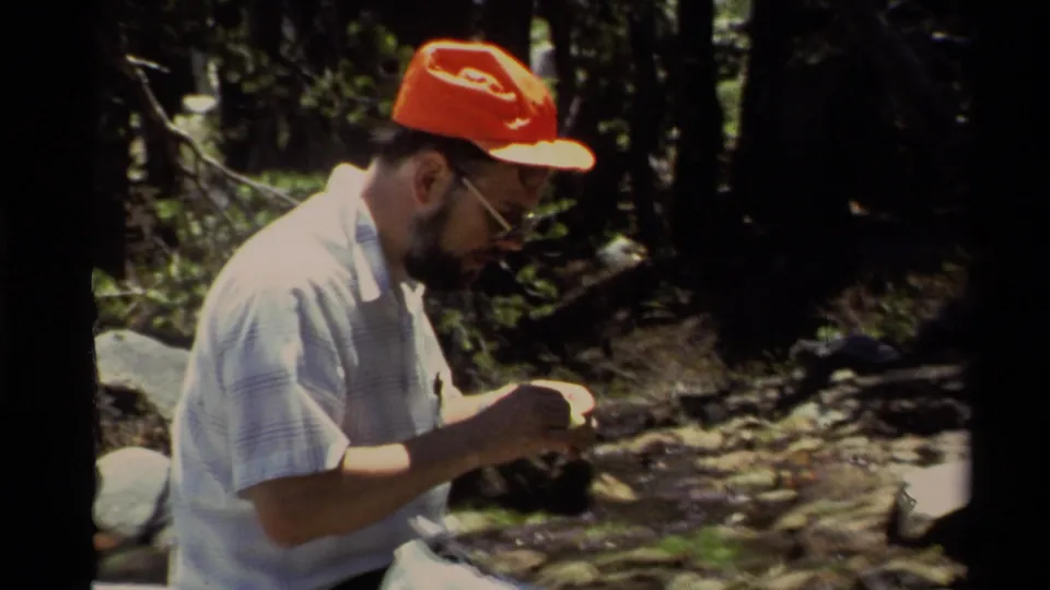 The man sits by a rocky creek or shoreline in 1978 Lake Tahoe footage, focused on fishing line or tackle in his hands.