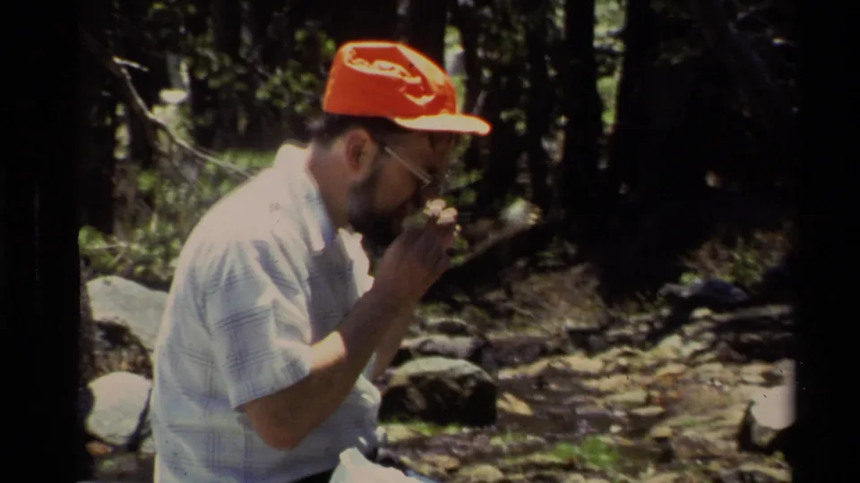 A man in a red cap and glasses appears to bite or adjust fishing line while seated in a wooded Lake Tahoe setting.