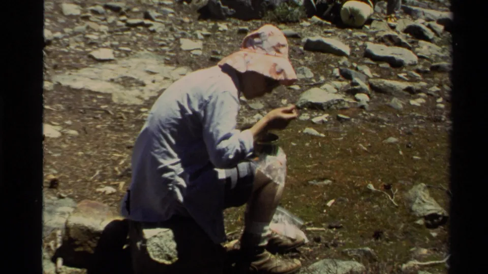 1978 Lake Tahoe 8mm frame of a hiker leaning forward with food in hand during a sunny backpacking rest.