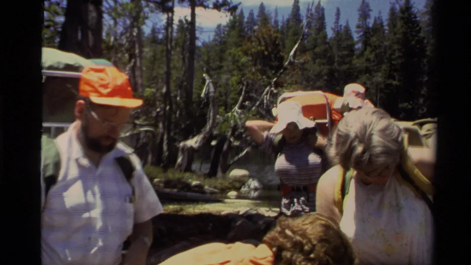 1978 Lake Tahoe 8mm frame showing adult backpackers adjusting gear beside a wooded lake shore.