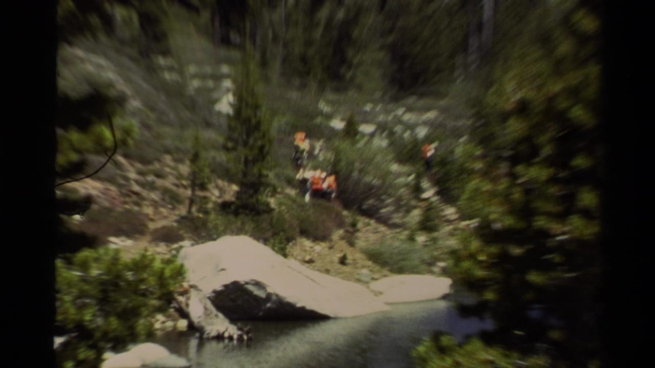 Handheld 8mm view at Lake Tahoe with distant hikers above a rocky shoreline, framed by pines and large pale boulders.