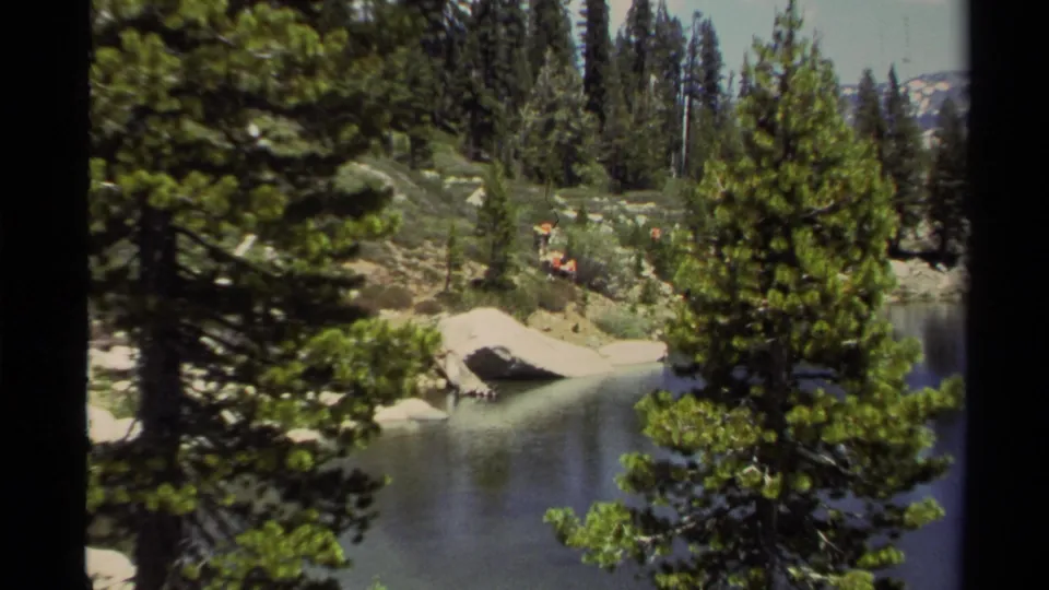 Lake Tahoe 8mm frame with evergreen trees in the foreground, calm water, boulders, and distant hikers along the shoreline area.