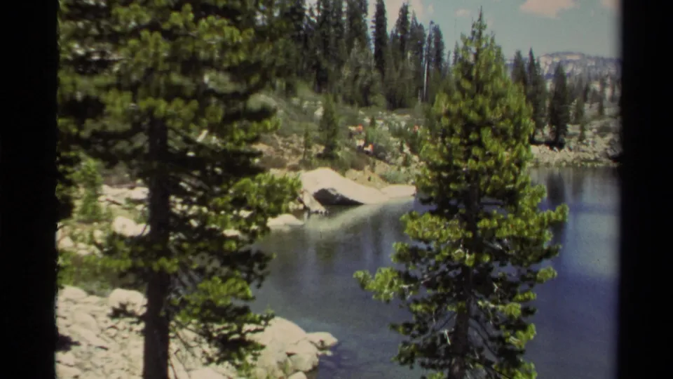 Wide 1978 Lake Tahoe home-movie view of pines and shoreline water, with hikers appearing small across the rocky lakeside terrain.
