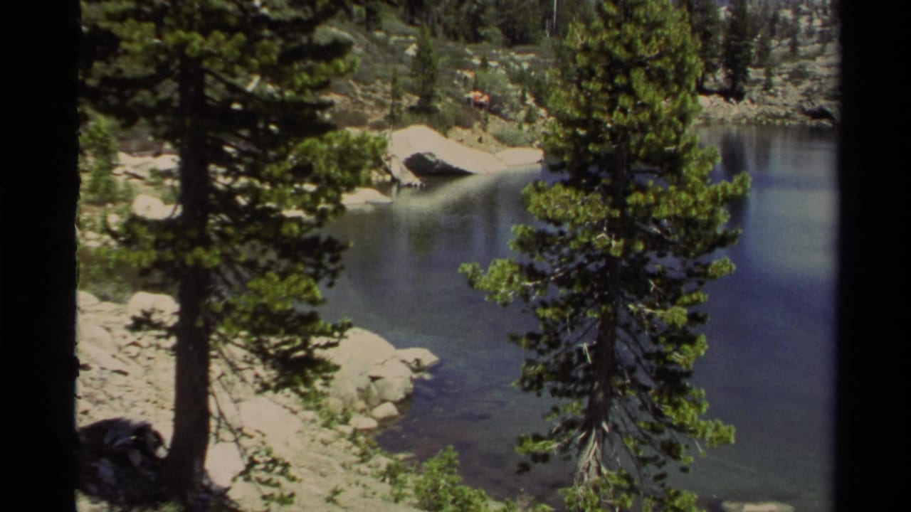 Lake Tahoe home-movie frame with conifers, pale shoreline rocks, and still lake water in the mountains.