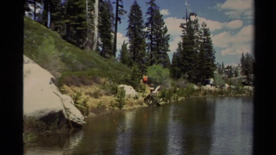 Lake Tahoe 8mm frame with a distant red-shirted hiker, tall pines, rocky bank, and calm water.