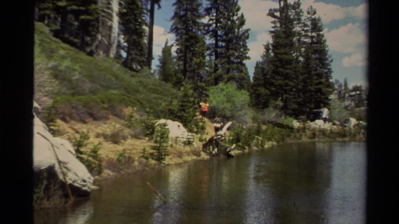 1978 Lake Tahoe home-movie frame showing a pine-lined lake or pond with a backpacker on a rocky forest trail.