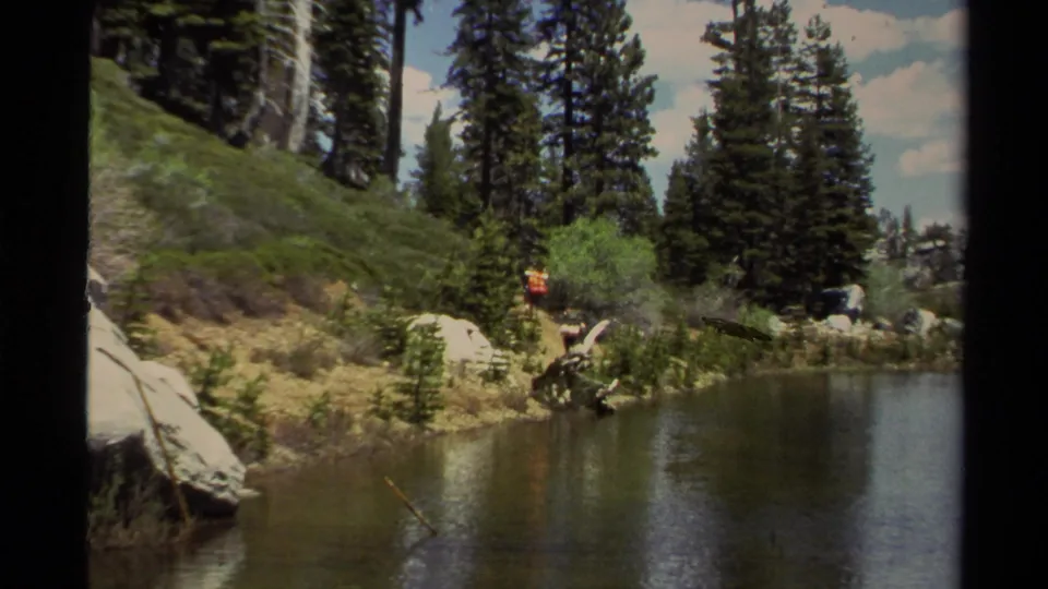1978 Lake Tahoe home-movie frame showing a pine-lined lake or pond with a backpacker on a rocky forest trail.