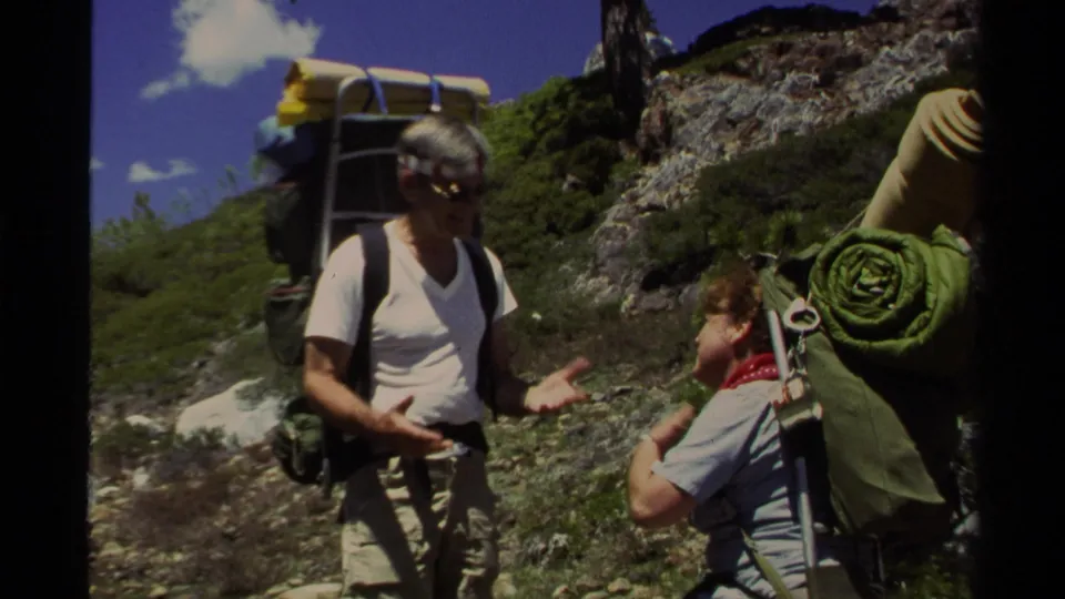 1978 Lake Tahoe home-movie frame of two hikers with large backpacks pausing on a rocky mountain trail under bright sky.