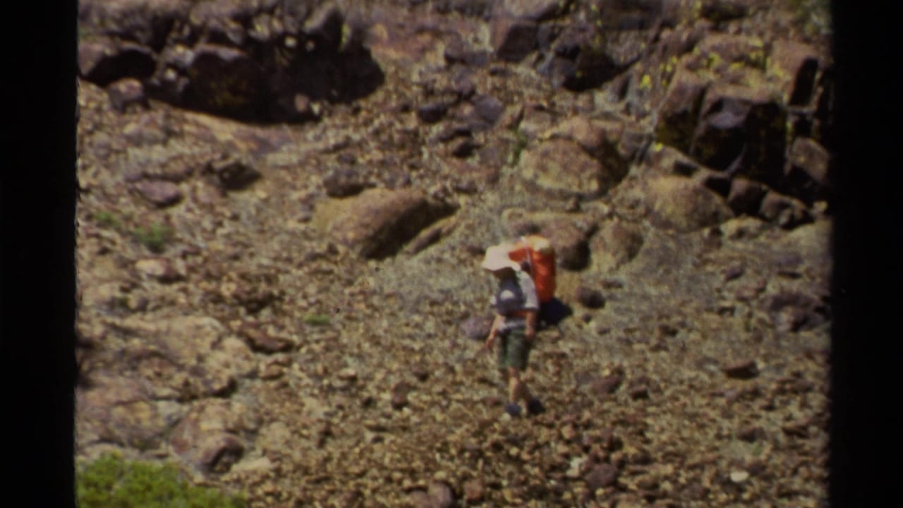 Distant hiker in 1978 Lake Tahoe footage steps across a rocky slope with a backpack and wide-brimmed hat.