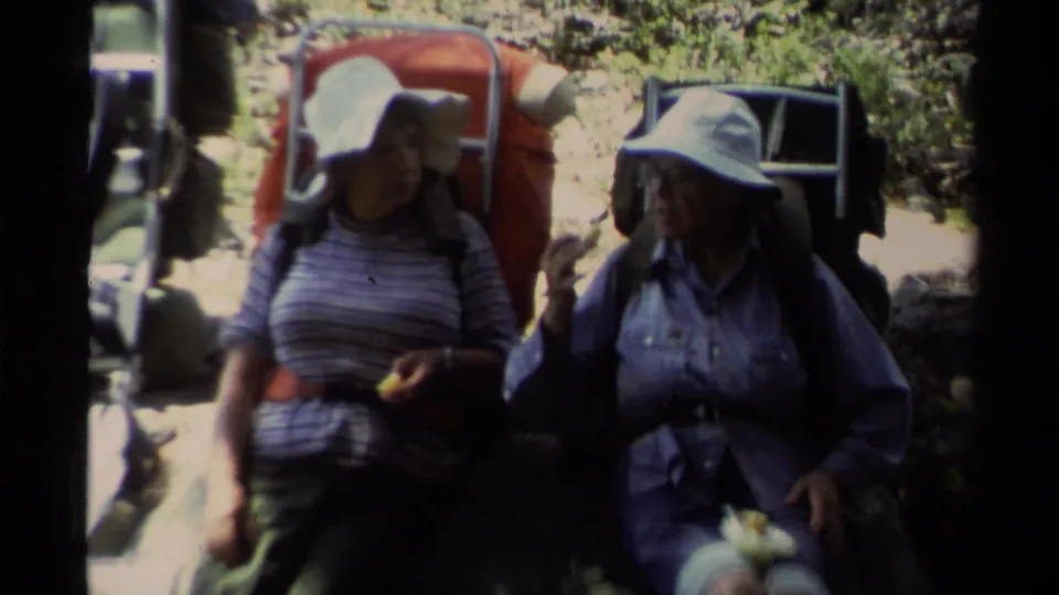 Two women hikers in wide-brim hats rest close to camera with large backpacks on a Lake Tahoe trail in 1978 home-movie footage.