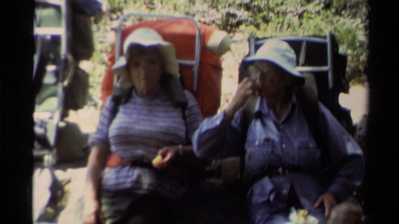 Two women with backpacks pause on a shaded forest trail, one eating and one drinking.