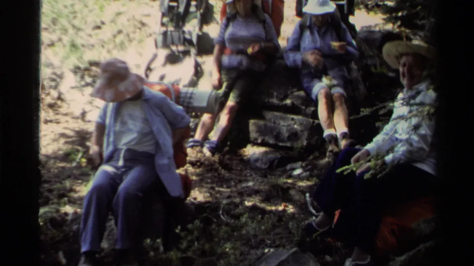 1978 Lake Tahoe home-movie frame showing hikers resting on rocks beside a wooded trail, with backpacks and sun hats visible.