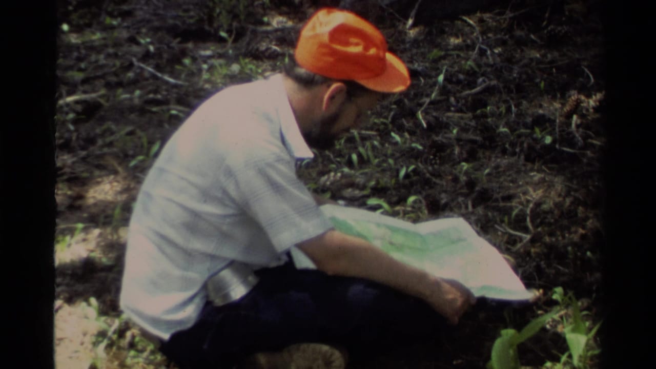 1978 Lake Tahoe home-movie frame of a man in an orange cap crouching near forest undergrowth while studying a paper map.
