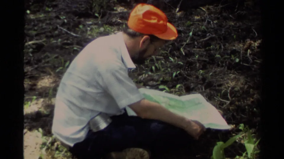 1978 Lake Tahoe home-movie frame of a man in an orange cap crouching near forest undergrowth while studying a paper map.