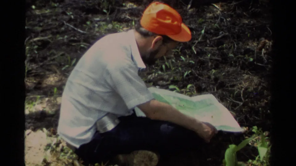 1978 Lake Tahoe home-movie frame showing a crouching hiker in an orange cap reading a map on a shaded forest trail.