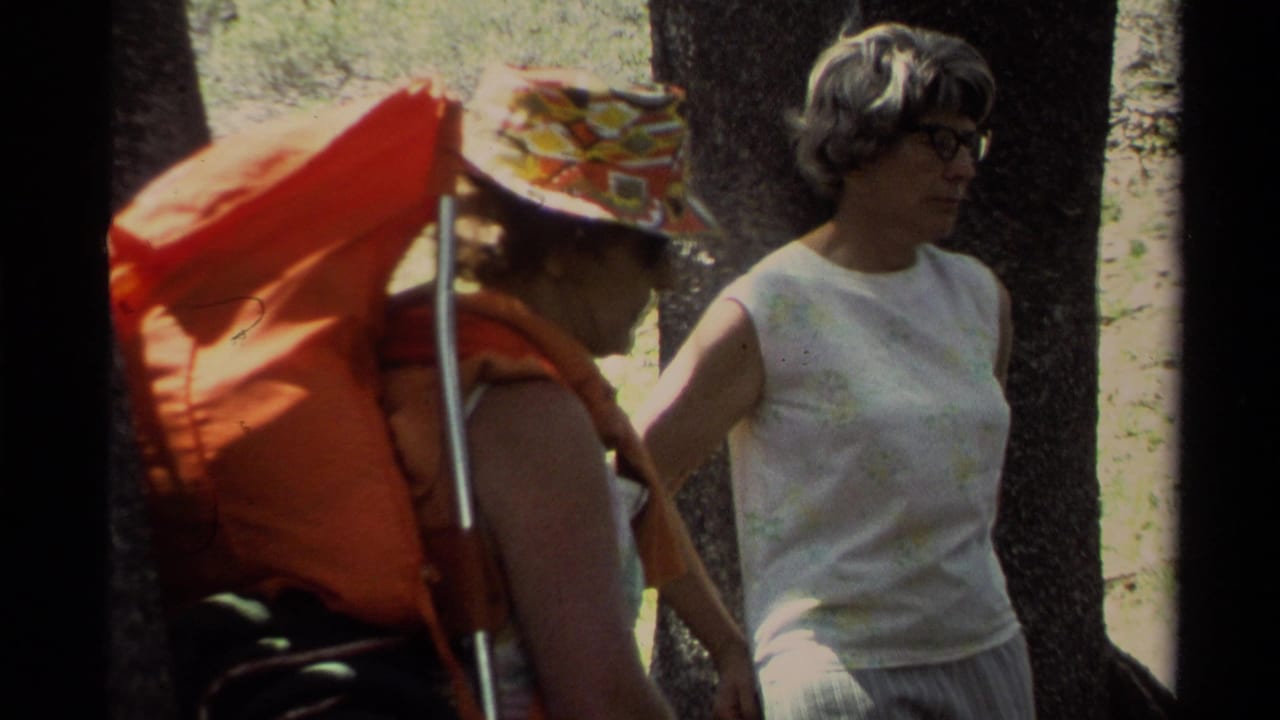 Two women stand beside tree trunks, with an orange backpack prominent on the left figure.