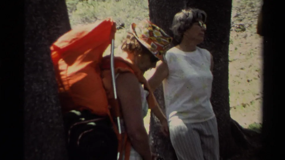A 1978 home-movie frame shows two adult women in wooded shade, one carrying a large orange camping pack.