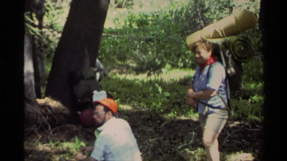 In the Lake Tahoe woods, an adult man squats near gear while an adult woman stands with a rolled sleeping bag on her backpack.