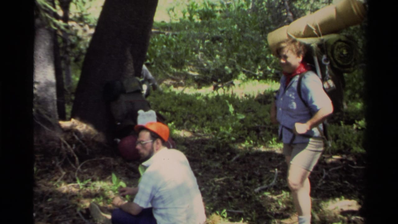 Two adult hikers rest in a wooded area, with a seated man in an orange cap and a standing woman carrying a bedroll.