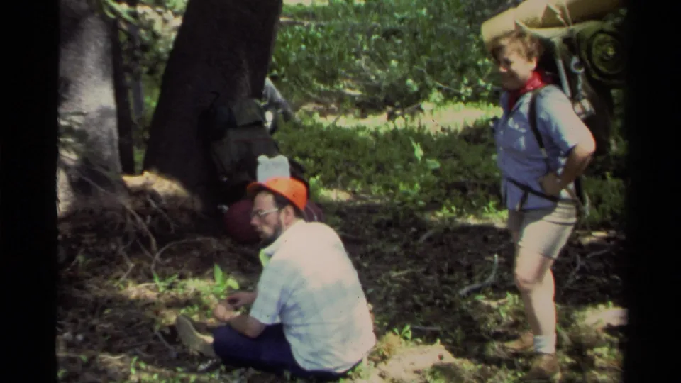 1978 Lake Tahoe forest scene with two adult hikers resting beside trees, carrying visible backpacking and camping equipment.