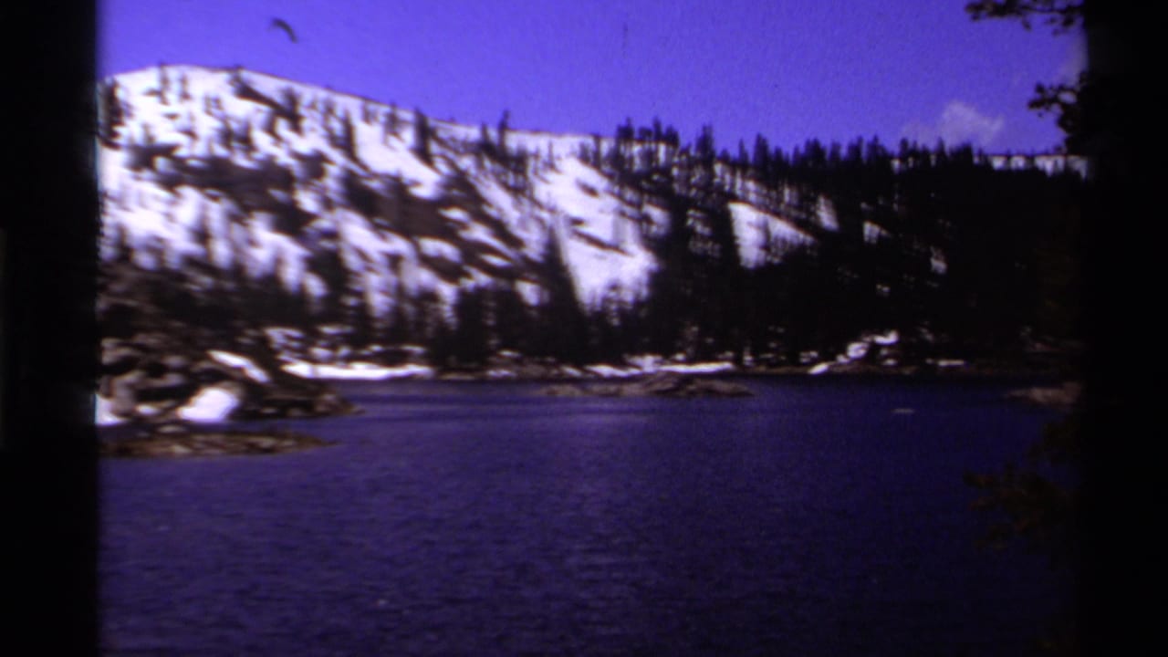 1978 Lake Tahoe frame with a dark blue lake in the foreground, snowy ridge beyond, evergreen trees, and shadowed film edges.