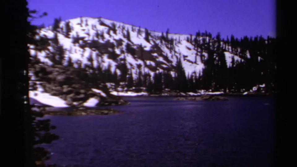1978 Lake Tahoe home-movie view of snow patches on a mountain slope above a forested shoreline and calm lake water.