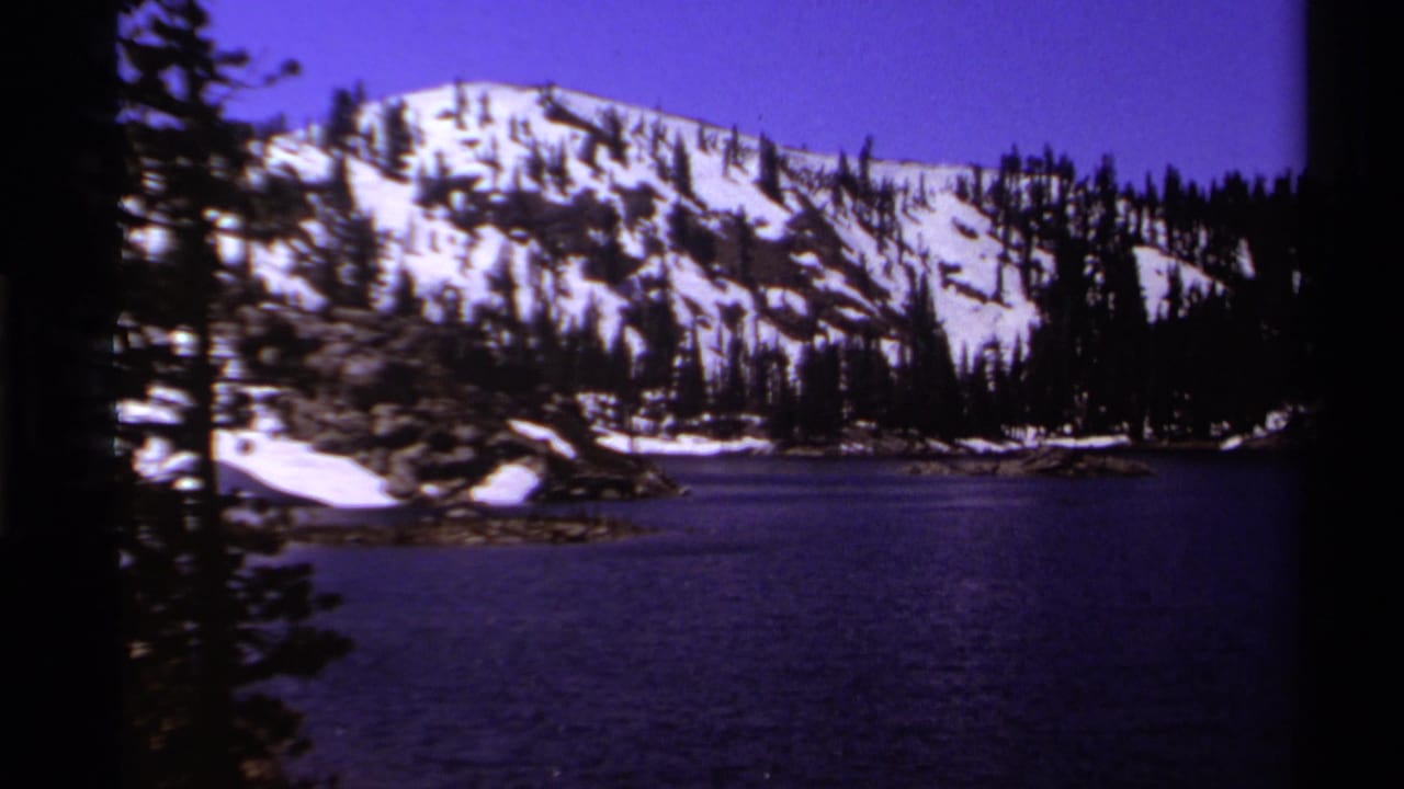 Mountain lake below a snowy rocky slope and pine forest, framed by dark film borders.