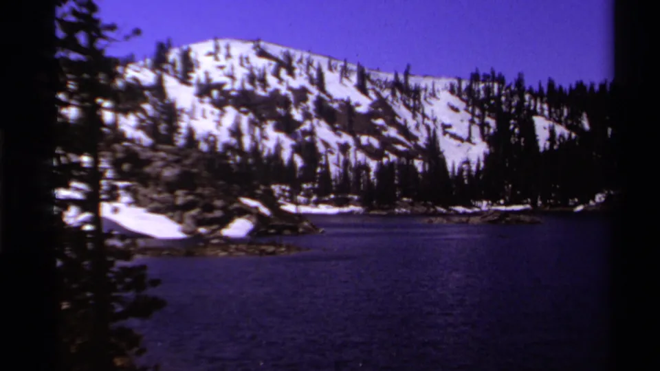 1978 Lake Tahoe home-movie frame showing a calm mountain lake, pine trees, snow patches, and rocky slopes under a blue-purple sky.