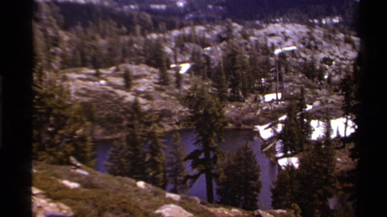 1978 Lake Tahoe frame looking down across rocks, evergreens, snow patches, and blue water in an alpine basin.