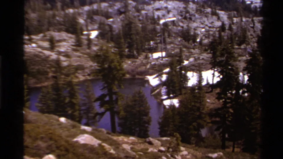 1978 Lake Tahoe home-movie view of evergreen trees, rocks, snow patches, and lake water during a slow landscape pan.