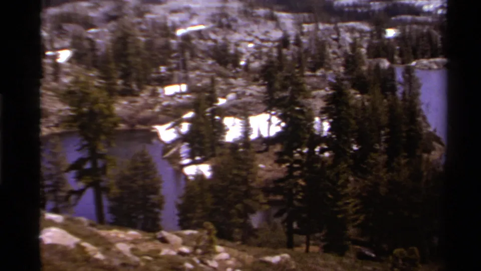 1978 Lake Tahoe frame showing pines, rocky lakeside slopes, bright snow patches, and blue water at the edge of the view.