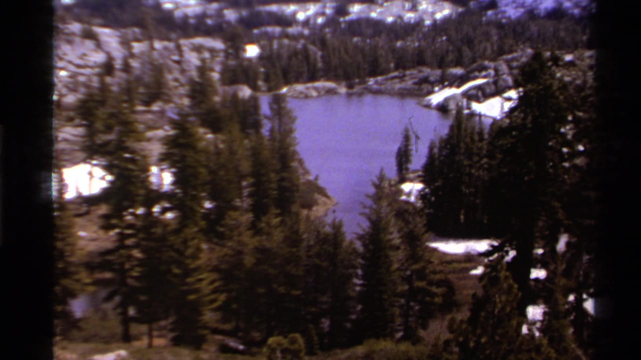 Lake Tahoe-region alpine lake in soft 8mm color, with evergreen forest, rocky shoreline, and patches of snow.