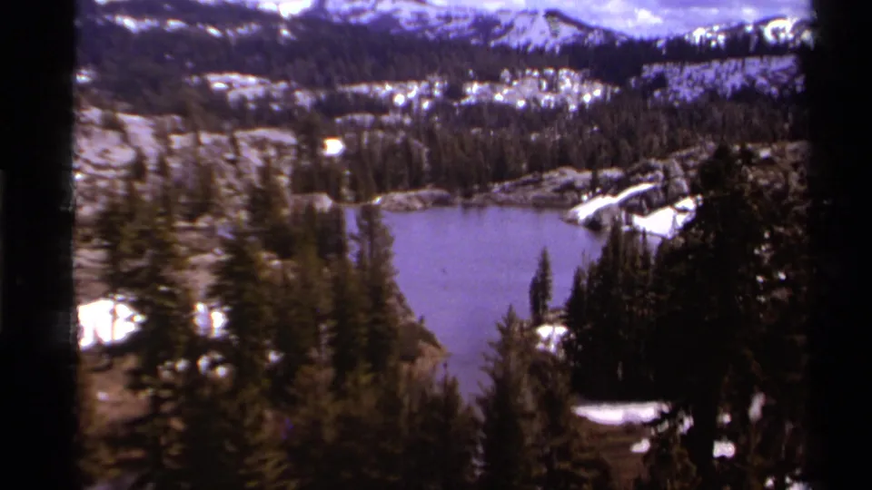 Wider 1978 8mm view of a Lake Tahoe-region alpine lake, forested slopes, snow, and distant ridgelines under cloud.