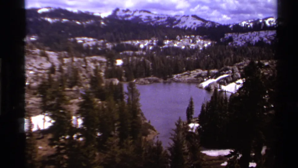 Elevated Lake Tahoe-region landscape on 8mm film, showing an alpine lake, evergreens, snow patches, and distant mountains.