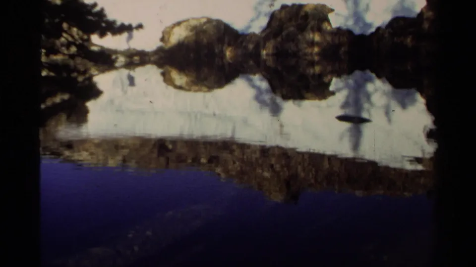 1978 Lake Tahoe 8mm frame with reflected clouds and shoreline shapes mirrored across a calm blue lake surface.
