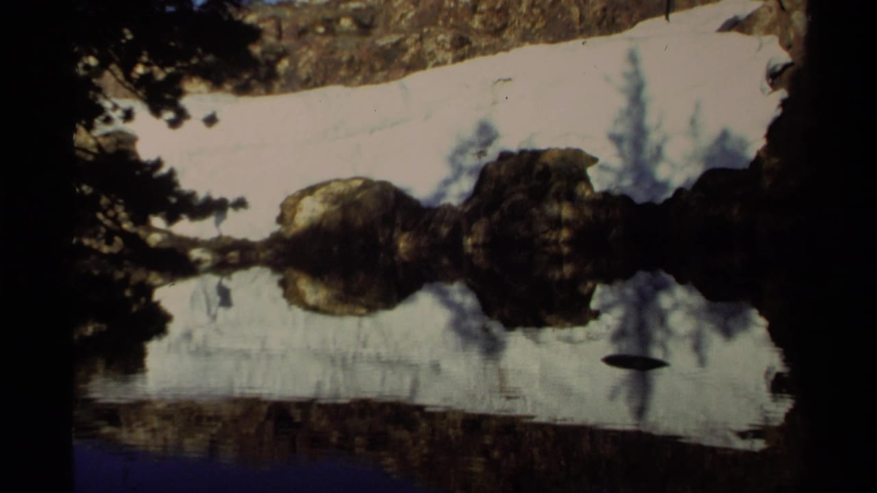 Lake Tahoe home-movie frame showing snow along the bank, rocks, evergreen shadows, and reflections on calm water.