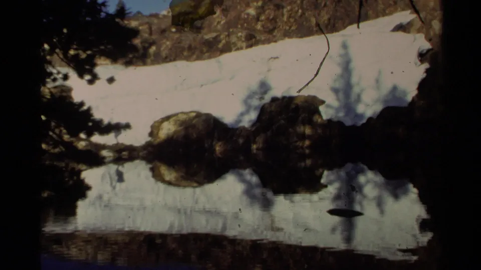 Archival 8mm view at Lake Tahoe with snow, shoreline rocks, evergreen silhouettes, and still water reflections.