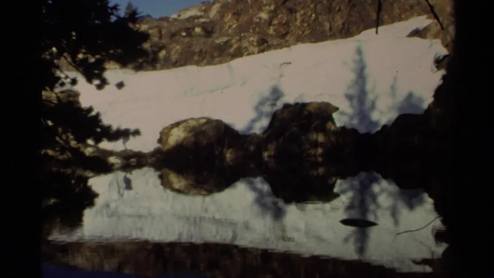 Lake Tahoe 1978 home-movie frame showing a snowy bank, rocky edge, evergreen shadows, and glassy water reflections.