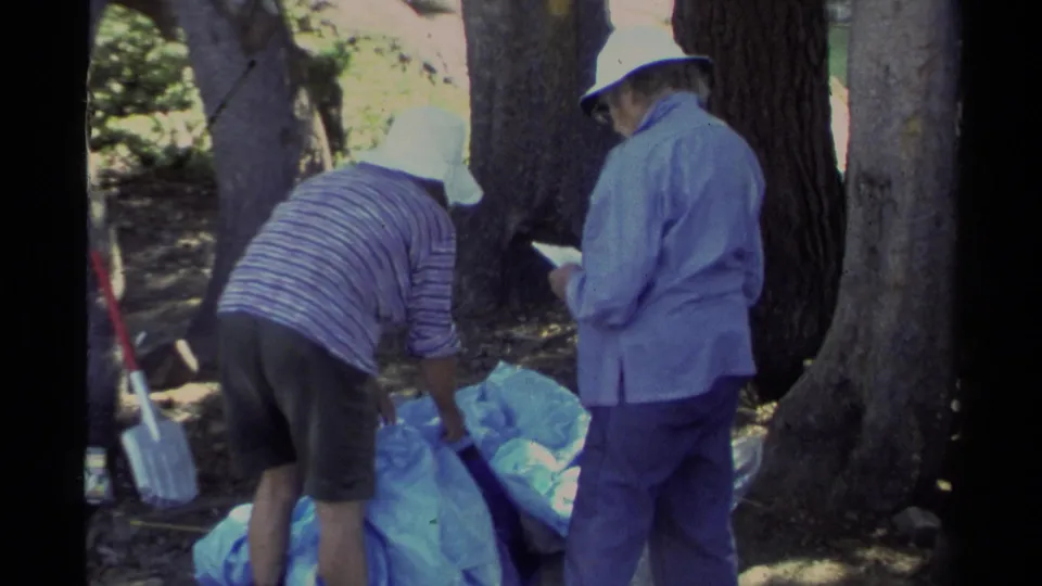 At a Lake Tahoe campsite, two adult campers in hats appear to collect trash or gear into a blue bag among trees.