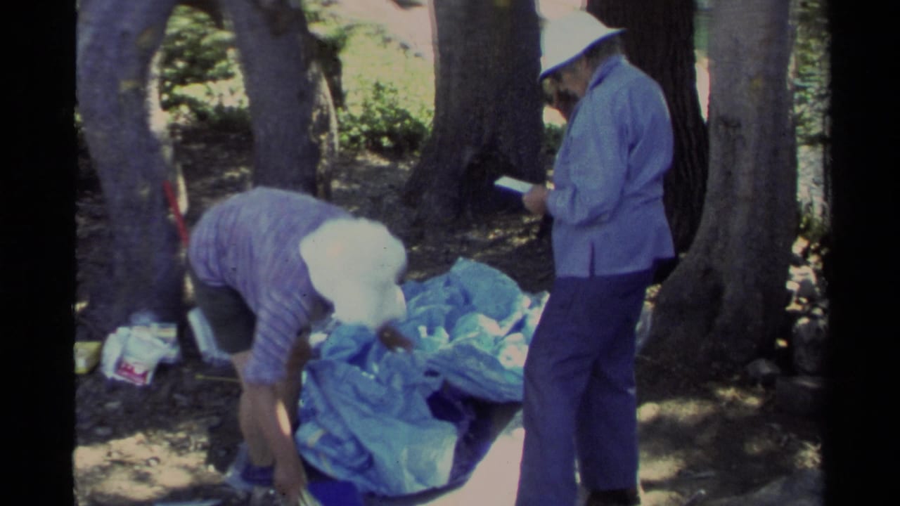 A Lake Tahoe camper bends toward blue tent material while a second camper in a light jacket consults a notebook in the woods.