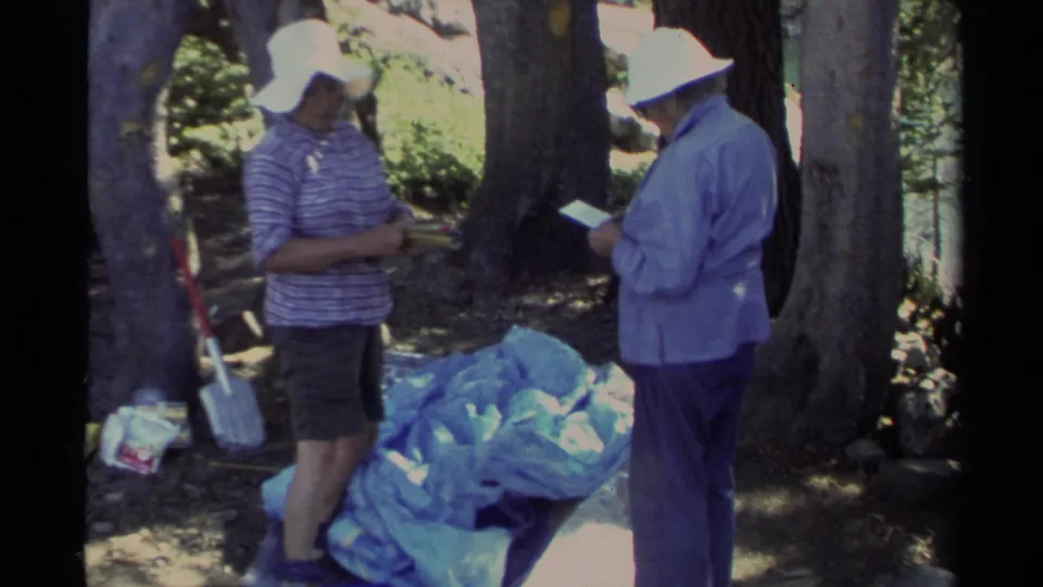 Two adult campers face each other near blue tarp or tent fabric in 1978 Lake Tahoe woods, with a shovel leaning behind them.