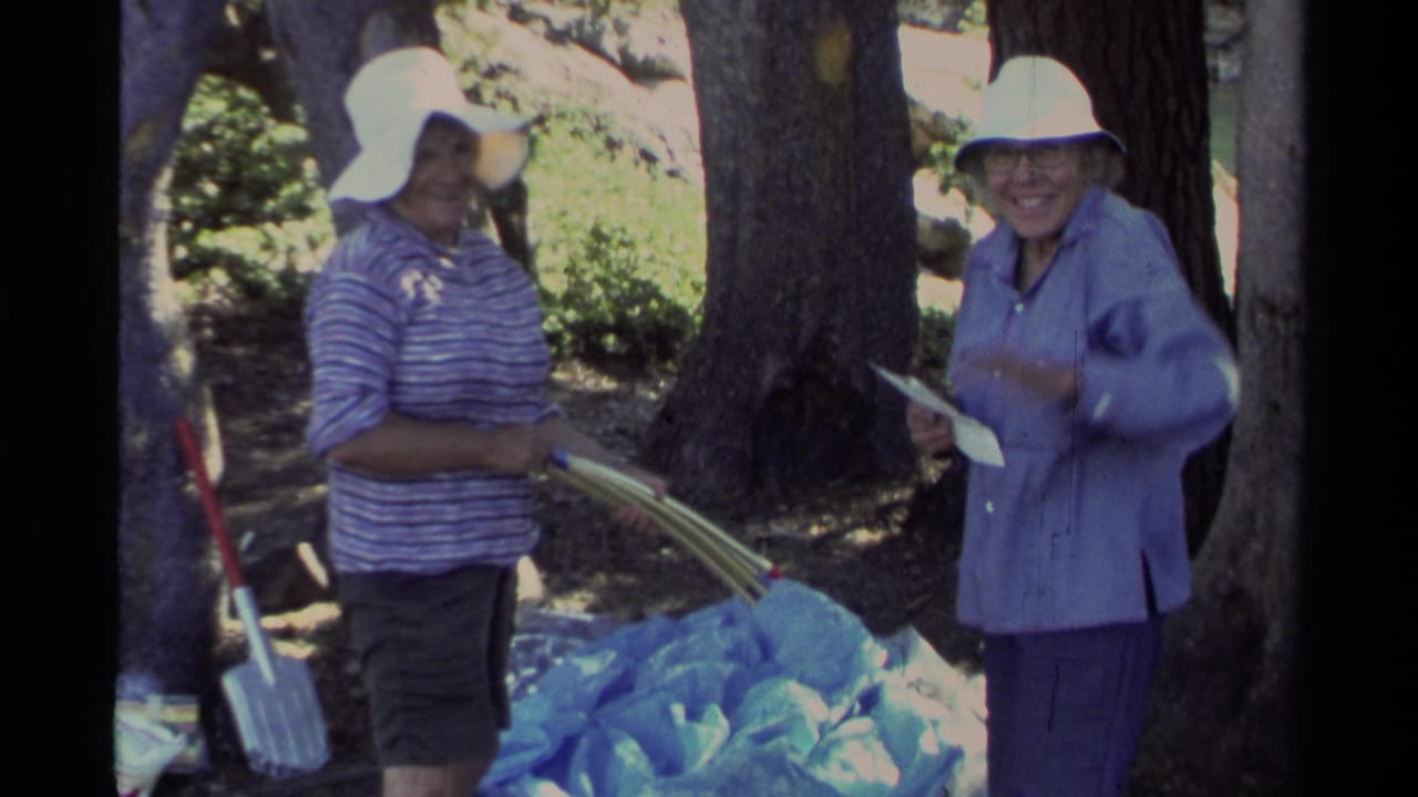 Two women in sun hats stand by blue camping fabric and poles under trees.