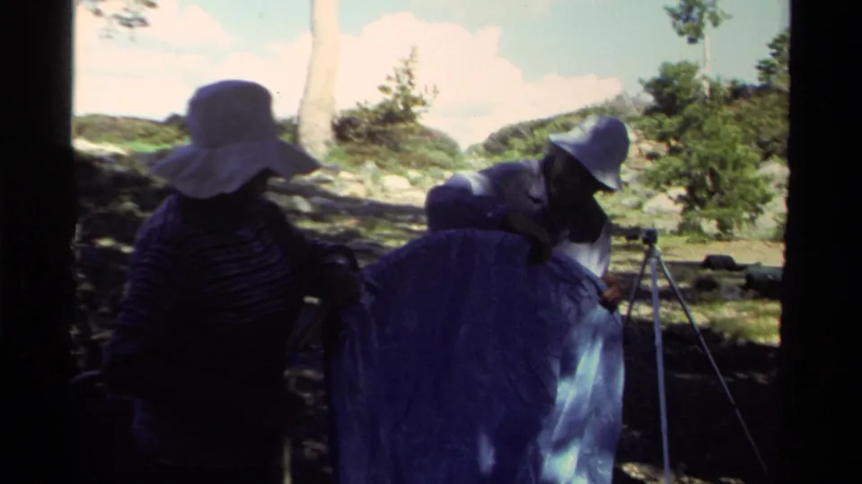 Two adults in sun hats lean over blue tent fabric while setting up camp near trees and a visible lakeshore at Lake Tahoe.