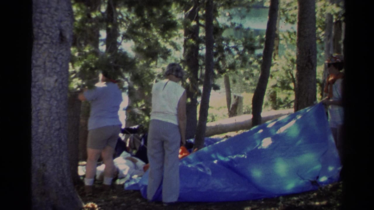 Adults in summer clothing adjust a bright blue tarp among Lake Tahoe trees, with camping gear nearby.