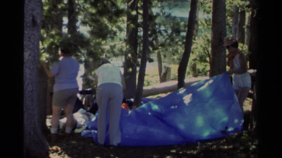Three adult women continue arranging a blue tarp and gear in a wooded Lake Tahoe campsite.