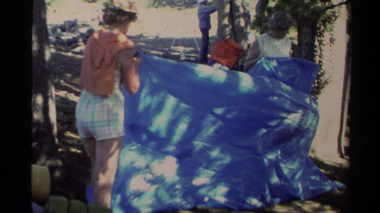 1978 Lake Tahoe home-movie frame showing adults working with a large blue tarp in dappled forest light.