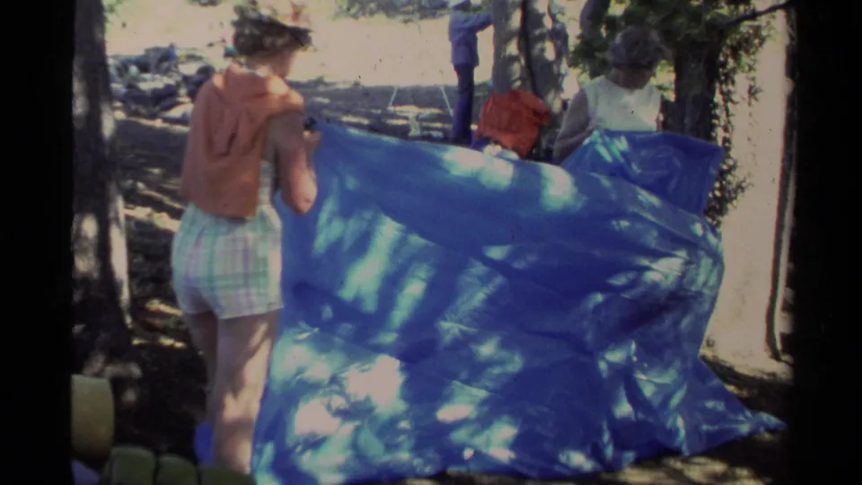 1978 Lake Tahoe home-movie frame showing adults working with a large blue tarp in dappled forest light.